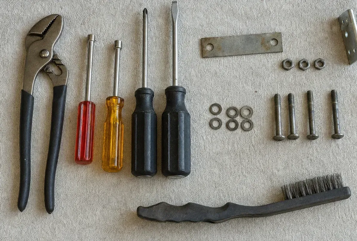 Slip-joint pliers and screwdrivers on canvas beside a flat strap plate, an angle bracket and a line of nuts, washers and bolts