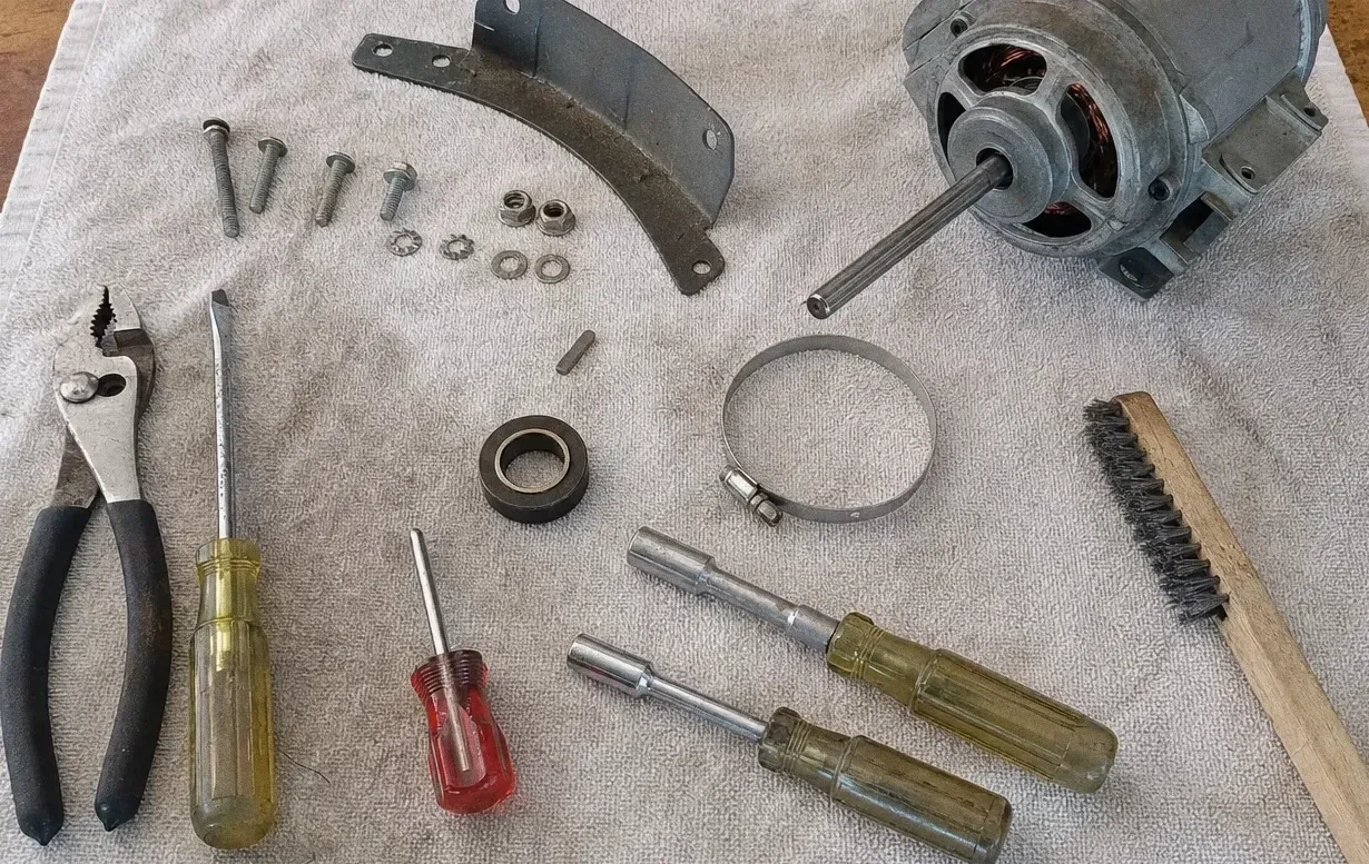 Small electric motor with its shaft, a hose clamp, a bearing and a bracket arm laid out on canvas with screwdrivers, pliers and washers